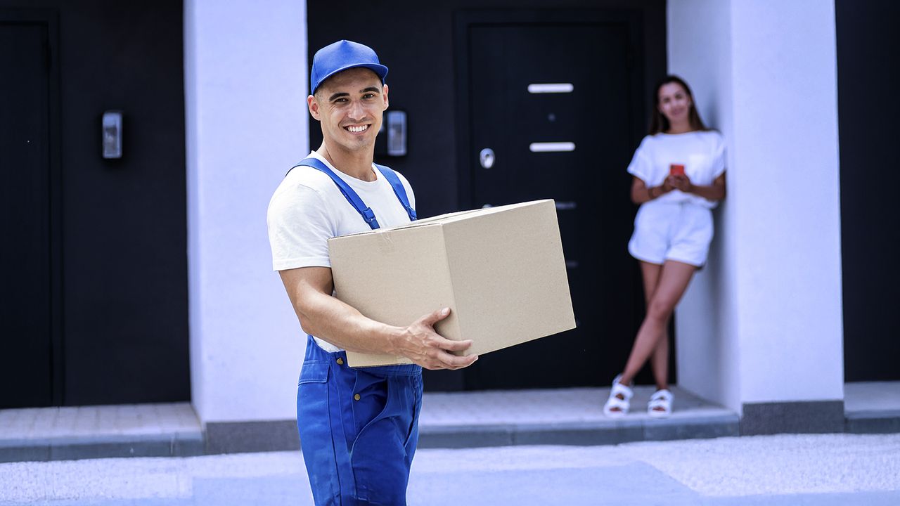 Transporte de carga con entrega directa al domicilio Repartidor sonriente entregando paquete frente a edificio residencial moderno